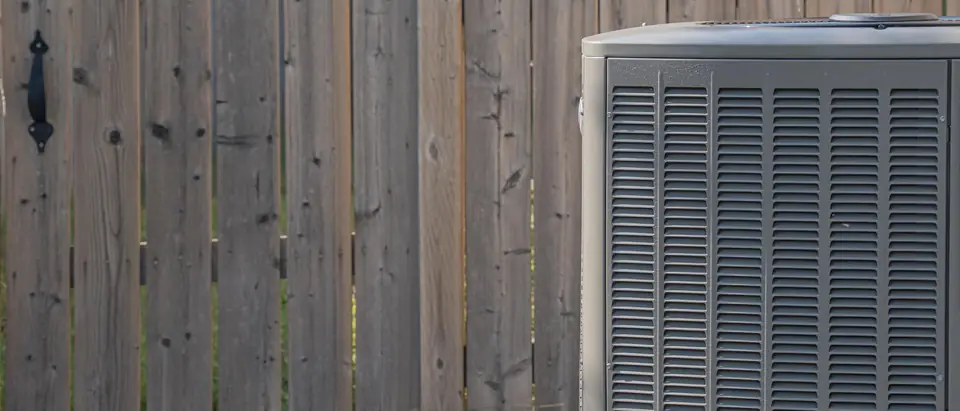 A gray outdoor condenser unit sitting on a concrete pad beside a weathered board fence