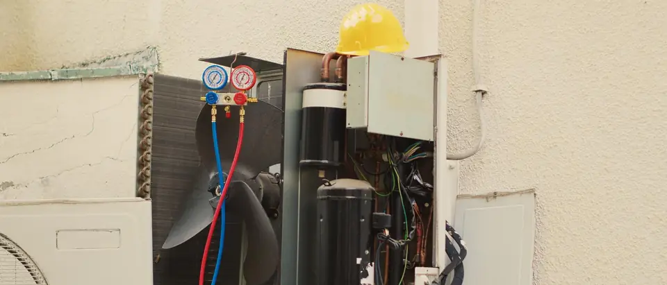 An outdoor air conditioner unit with its panels off against a stucco wall, showing the fan, compressor and wiring, with a set of refrigerant gauges hung on it