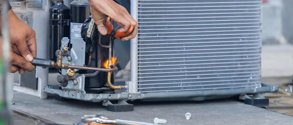 Two hands brazing a copper refrigerant line beside the compressor of an opened outdoor unit, one holding a torch and the other pliers, with tools on the ground