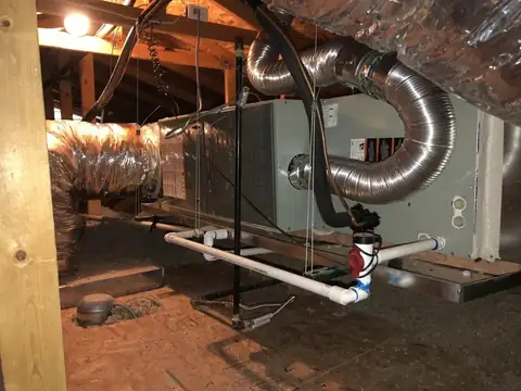 An air handler hung in a house attic with flexible ducts and a white condensate drain, above a floor of loose-fill insulation and a plywood service deck