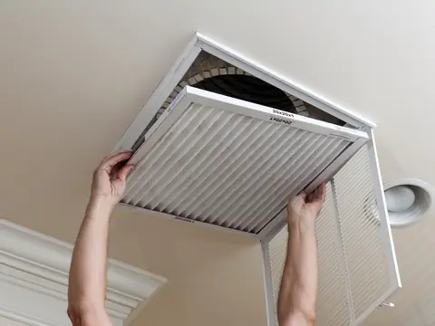 Two hands sliding a pleated air filter into an open return air grille in a ceiling
