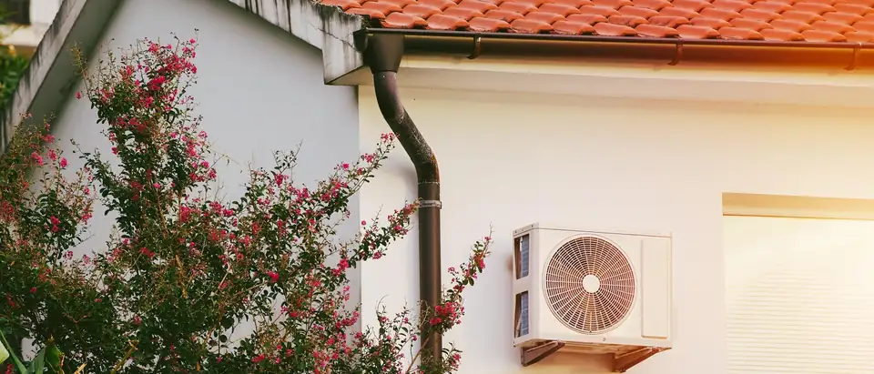 The outdoor unit of a ductless heat pump mounted on the white stucco wall of a house with a red clay tile roof, beside a shrub in pink flower