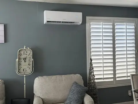 A ductless mini split indoor unit mounted high on a blue-gray living room wall beside a window with white shutters