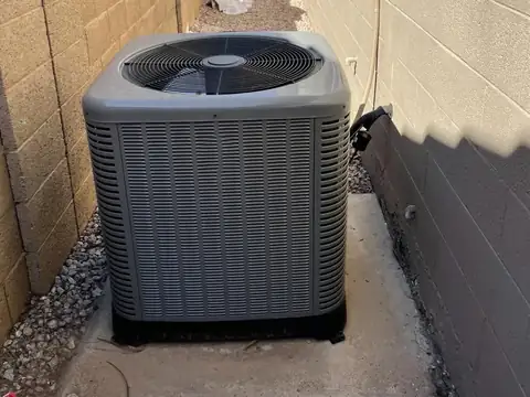 A new gray condenser unit set level on a concrete pad in a gravel side yard, between a tan block wall and the block wall of a house
