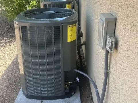 Two new condenser units on a concrete pad beside the stucco wall of a house, with their electrical disconnects on the wall