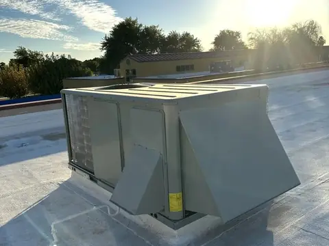 A package air conditioning unit with its duct hoods on a flat coated roof, with the sun low over trees behind it