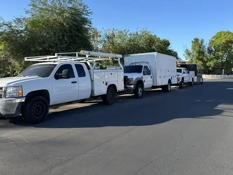 Three white service trucks, one with a ladder rack and one towing a black enclosed trailer, parked along a residential street under a clear blue sky